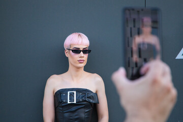 Young gay boy with pink hair and make-up and dressed in pink and black is leaning against a black...