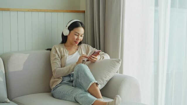Happy young Asian woman with mobile phone listening music in headphones and relaxing on safa at home