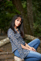 A confident, stylish woman in jeans and blue blouse poses casually in a sunny park, exuding modern urban elegance
