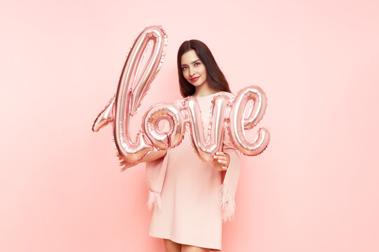 A beautiful young woman holding a balloon in the shape of an inscription of love on a pink background on Valentine's Day. The symbol of love