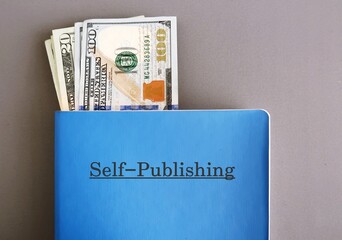 Cash dollars money in blue notebook with text typed SELF - PUBLISHING, on grey background, concept of make more income from writing book or ebook and self-publish online or traditional way