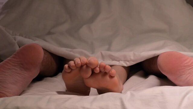 legs of a couple having sex under a blanket on a white sheet on a bed in a hotel bedroom. A man and a woman are making love