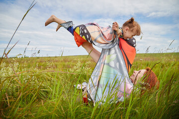Happy female family with mother and daughter on green and yellow meadow full of grass and flower. Woman with red hair and blonde girl having fun, joy and hugs in sunny summer day. Concept family love