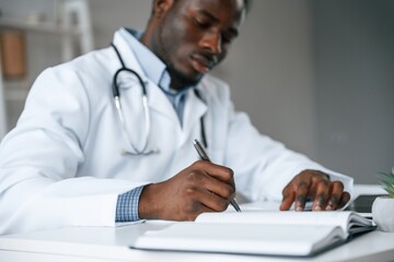 Filling in the document, writing by using pen. Black man as doctor in the clinic office