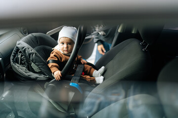 View from side window to cute infant baby boy sitting on front seat with buckled security belt in...