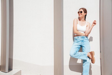 Fototapeta na wymiar Beautiful smiling model in sunglasses. Female dressed in summer hipster white T-shirt and jeans. Posing near white wall in the street. Funny and positive woman having fun outdoors, in sunglasses