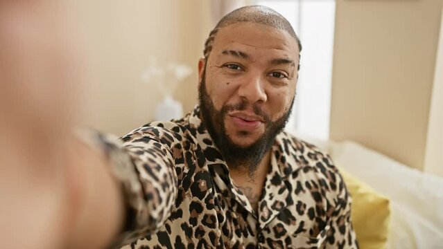 African american man with beard in leopard print shirt smiling in a bedroom setting, taking a selfie.