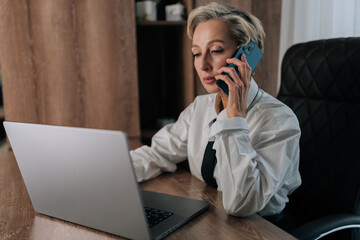 Successful serious middle-aged business woman talking on smartphone siting at office desk with laptop, share helpful information, solve business, lead formal conversation to client working at office.