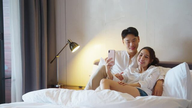 Asia people young adult man woman relax hug smile look camera take photo record video post to social media at hotel cozy bed enjoy date day. Happy asian lover wife husband just married sweet newlywed