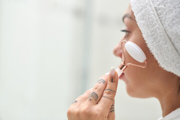 Young woman with soft white towel on head having procedure of facial massage with jade stone roller after morning shower in bathroom - obrazy, fototapety, plakaty