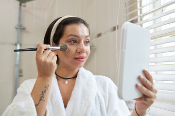 Young brunette woman in bathrobe looking in mirror in her hand while applying loose powder or blush...