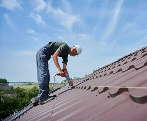 Worker prepearing to installation photovoltaic solar panel system on the roof of house. Man installer working with electric screwdriver, wearing uniform and helmet, on blue sky background.