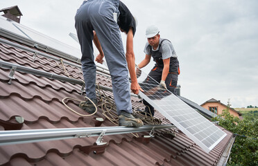 Men technicians lifting up photovoltaic solar moduls on roof of house. Workmen in helmets mounting solar panel system outdoors. Concept of alternative and renewable energy.