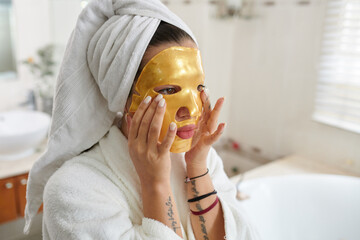 Young woman in bathrobe with towel on head putting golden rejuvenating mask on her face while...