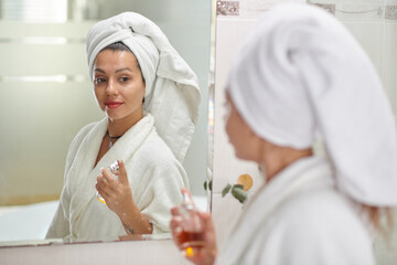 Young woman with towel on head standing in front of mirror in bathroom and spraying fragrance from...
