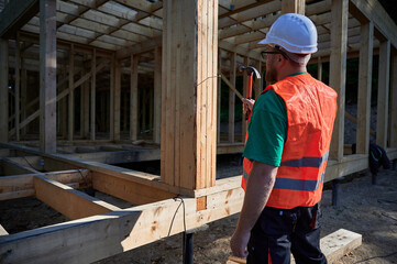 Carpenter constructing two-story wooden frame house near the forest. Back view of man hammering nails into structure while wearing protective helmet and construction vest.