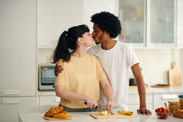 Two young intercultural sweethearts kissing one another while affectionate woman chopping fresh...