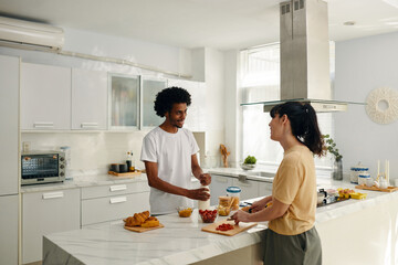 Happy young husband and wife in t-shirts standing by kitchen table and looking at one another while woman chopping tomatoes for salad