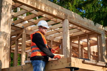 Carpenter constructing wooden frame house near forest. Bearded man with glasses hammering nails...