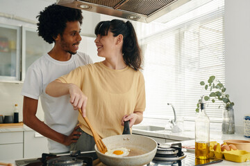 Pretty young woman with frying pan cooking scrambled eggs for breakfast while standing by electric stove and looking at her husband - obrazy, fototapety, plakaty