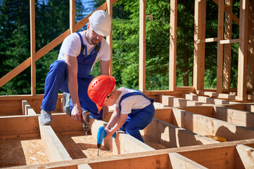 Father with toddler son constructing wooden frame house. Male builder coaching his son on how to operate an electric screwdriver on construction site on sunny day. Carpentry and family concept.