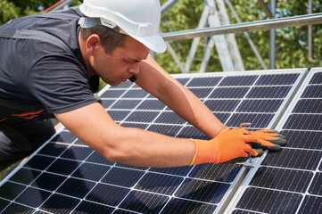 Male worker mounting photovoltaic solar panel system outdoors. Man engineer placing solar module on metal rails, wearing construction helmets and work gloves. Renewable and ecological energy.