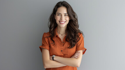 Fototapeta premium a cheerful young woman with long wavy hair, wearing an orange blouse, standing with her arms crossed against a neutral gray background