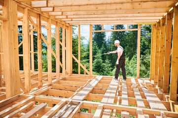 Carpenter is constructing wooden frame house. Man taking measurements using tape measure while...