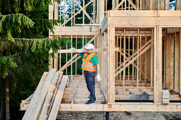 Carpenter building wooden frame house near the forest. Man holds large truss on his shoulder, dressed in work clothes and helmet. Concept of contemporary and sustainable construction.