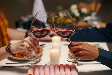 Hands of young unrecognizable sweethearts clinking with glasses of wine over table served with homemade food and fresh platter