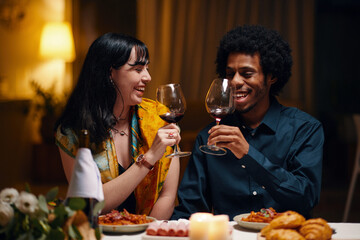 Happy young man and woman with glasses of red wine toasting for love and Valentine Day while sitting by served table at dinner