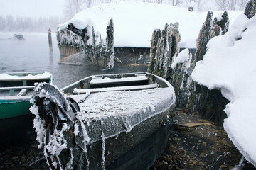 Snow-covered wooden boat near river shore in winter. Old fishing and rowing boats with snowy hulls.