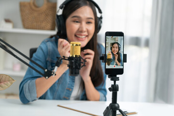 Fototapeta na wymiar Host channel Asian influencer talking in broadcast wearing headsets on social media live on smartphone recording online, greeting listeners with coaching life or business at modern studio. Stratagem.