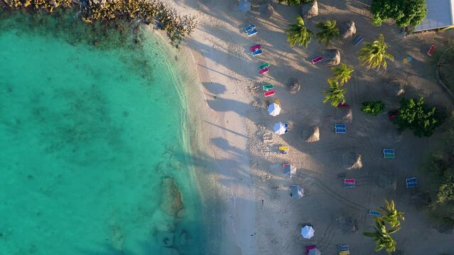 Drone descends on long shadows from palm tree and umbrellas on hidden tropical cove beach of Daaibooi Curacao