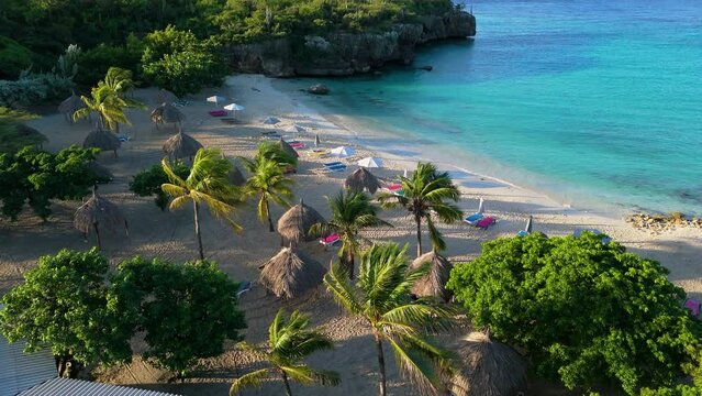 Aerial dolly above tropical beach side huts and palm trees with long shadows, ocean waves crash on Daaibooi beach