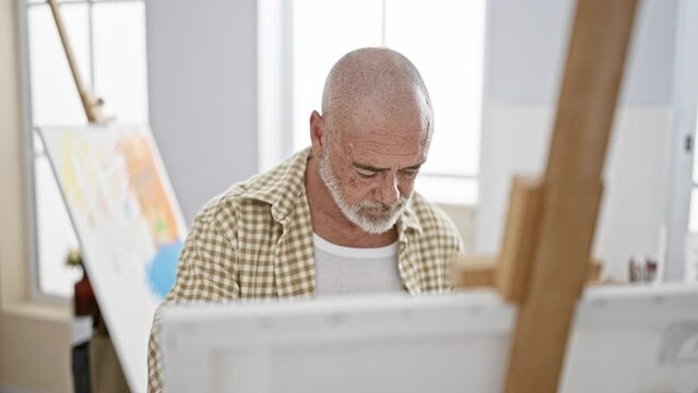 A senior man with a beard focusing intently on his artwork in a bright art studio