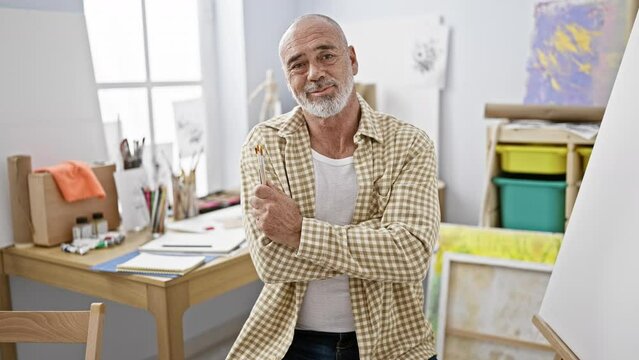Confident senior man with grey beard in art studio holding paintbrushes, creativity concept.