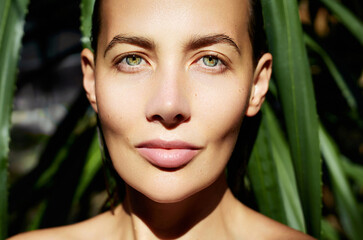 Skin hydration. Natural organic skincare. A close-up portrait  of a woman's face bathed in sunlight, with green leaves in the background, highlighting her natural beauty and striking green eyes.