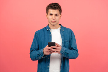 Portrait of serious man using mobile phone, texting on pink background looking at camera. Concept of human emotions, facial expression. Copy space for your ad