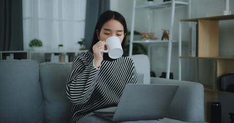 Portrait of Happy young asian woman sit on sofa drinking coffee or tea while working creative job from home on laptop with online network,take break,smiling