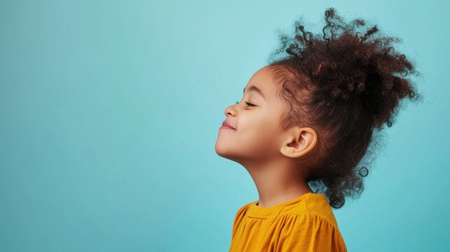 A young child with curly hair wearing a yellow top looking up with a smile against a light blue background.