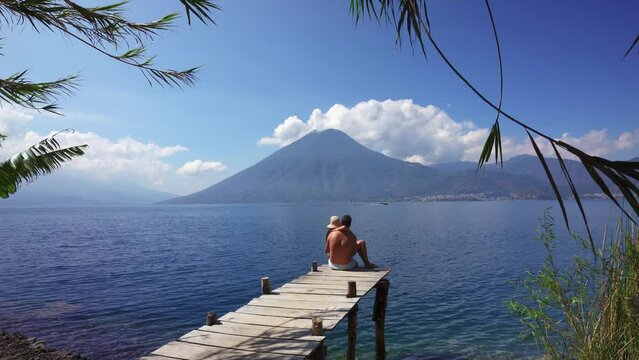 Man and woman sitting on pier watching boats on the lake go past a volcano in Lake Atilan, Guatemala