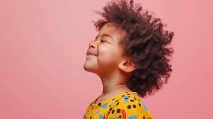 A joyful young child with curly hair wearing a yellow shirt with a playful pattern looking up towards the sky with a smile on their face set against a soft pink background.