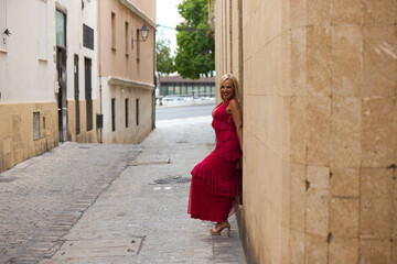 Fototapeta na wymiar Beautiful blonde mature woman in an elegant dress leaning against the wall of a building on a narrow street in a town in Andalusia, Spain.