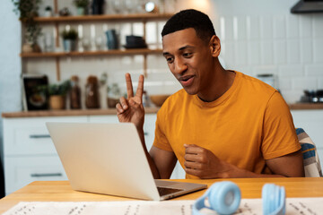Portrait of attractive young African American man wearing stylish t shirt, using laptop, sitting in kitchen in modern apartment, showing victory sign, having video call. Online communication concept - obrazy, fototapety, plakaty