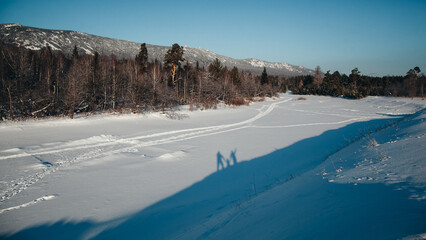 Couple walking on frozen lake, snow-covered trees, and distant mountains under clear blue sky