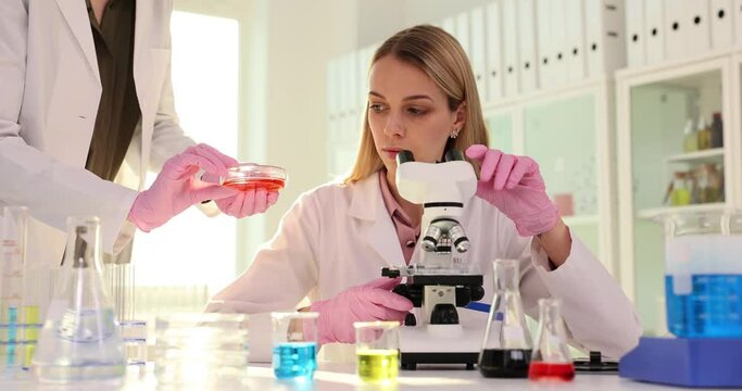 Female scientist works with microscope and a colleague gives test tube for analysis in modern laboratory