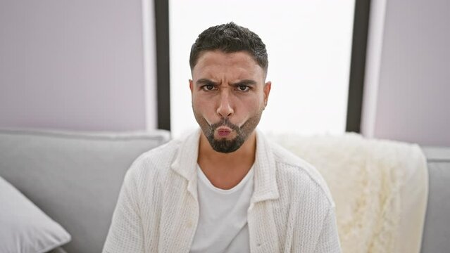 Cheeky young arab man sitting on living room sofa, making a crazy fun fish face and comical lips gesture. hilarious expression at home!