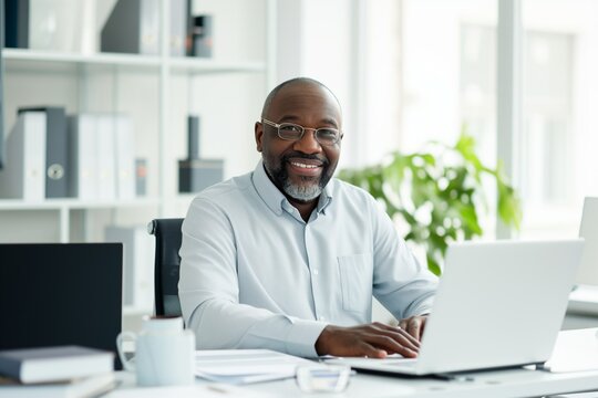 A smiling, contented black businessman in a shirt is working at an office desk with a laptop