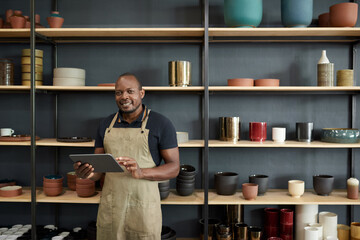 African potter with a tablet smiling by shelves in a ceramics workshop